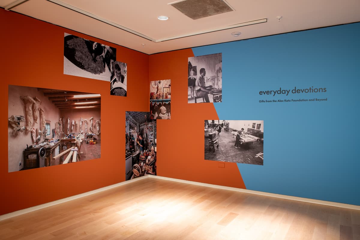 A corner view of the gallery where a blue wall meets a rust-orange wall. Various photographs of interior studio spaces and artists at work are displayed across both colored surfaces.