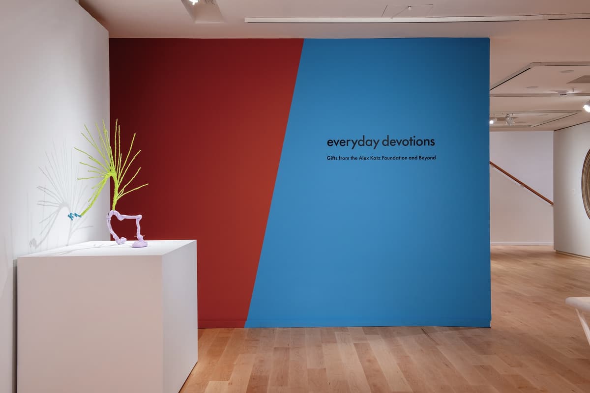 A bright blue and rust-red accent wall featuring the exhibition title "everyday devotions." To the left, a small, spindly neon-yellow and purple sculpture sits on a white pedestal.