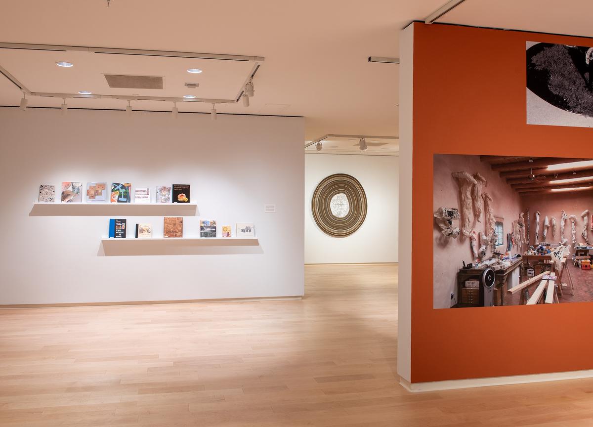 View from a doorway showing a white gallery wall with two narrow floating shelves holding various art books. To the right, a rust-orange wall features a large photograph of a studio.