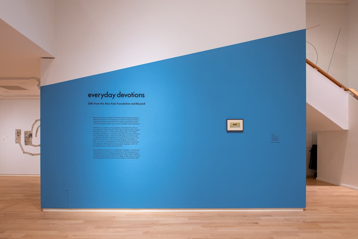 Full view of a blue accent wall with a diagonal top edge. The exhibition title and introductory text are printed in black on the left, next to a small framed artwork.