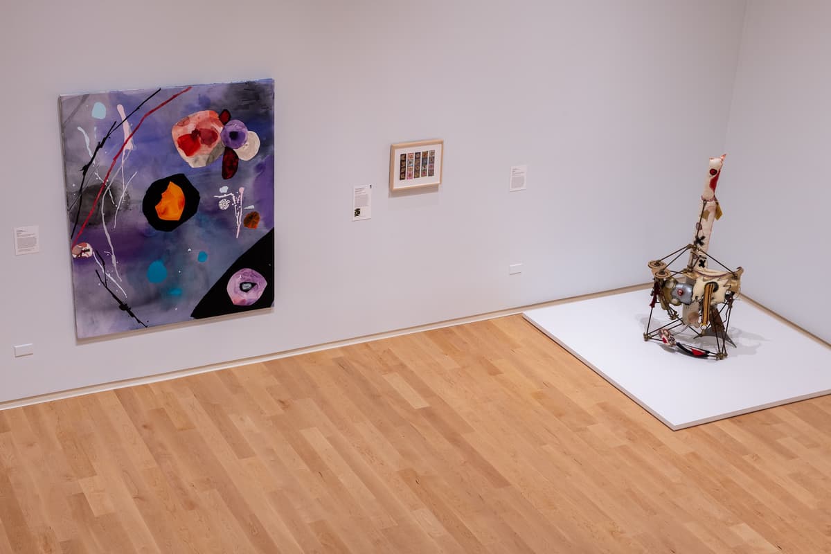 Close-up of a white wall featuring two framed abstract collages alongside a larger, brightly colored painting with blue, green, and orange geometric shapes and black dots.