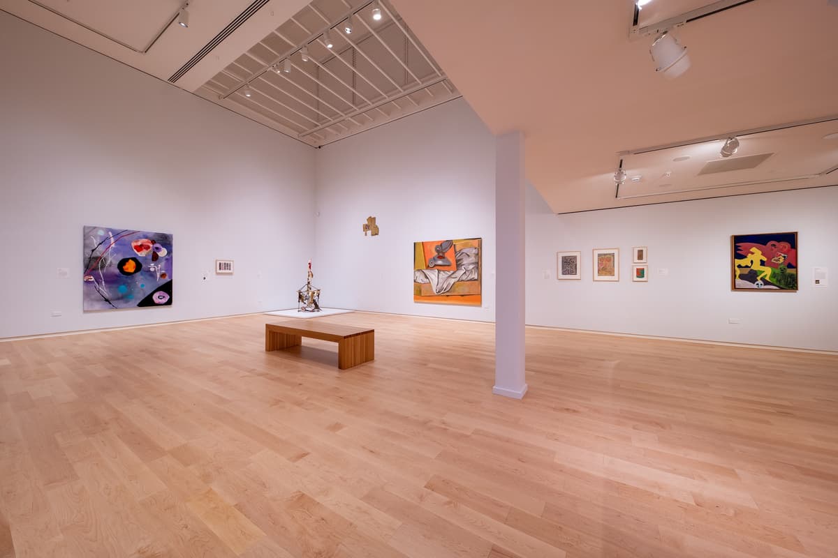 An installation view showing a white wall with several small framed works. In the foreground, a tall, thin neon-yellow and lavender sculpture stands on a white pedestal.