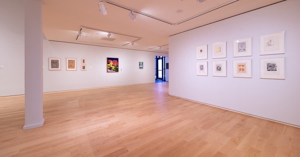 Gallery view of a long white wall with several small framed artworks and a colorful quilt. A doorway at the end leads to another gallery space with a large circular fabric sculpture.