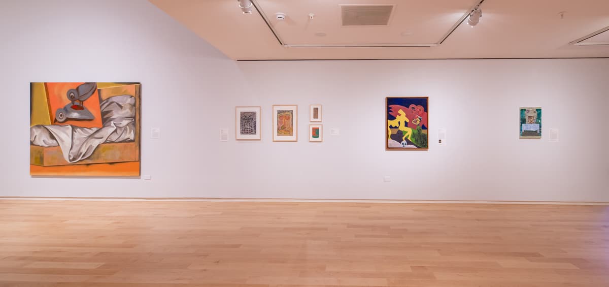 A wide shot of the gallery showing various small framed works on a white wall. The composition leads the eye toward a distant white pedestal holding a colorful, spindly sculpture.