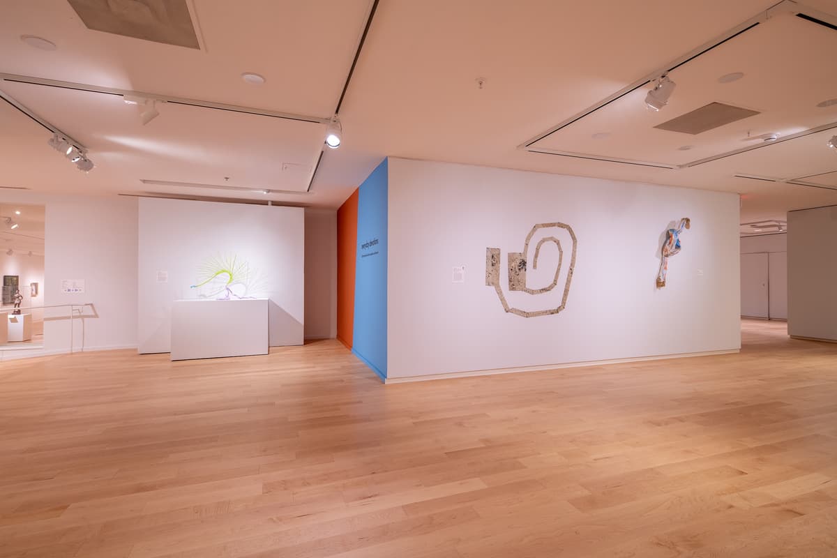 A corner view of the gallery featuring three framed works on paper and a small, dark, textured wall-mounted sculpture. A blue accent wall is visible in the background.