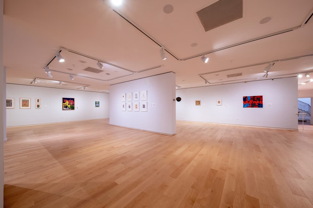 A long perspective of the gallery showing a series of small artworks on the left wall, leading toward a brightly lit area with a sculpture and a large blue and red mural.