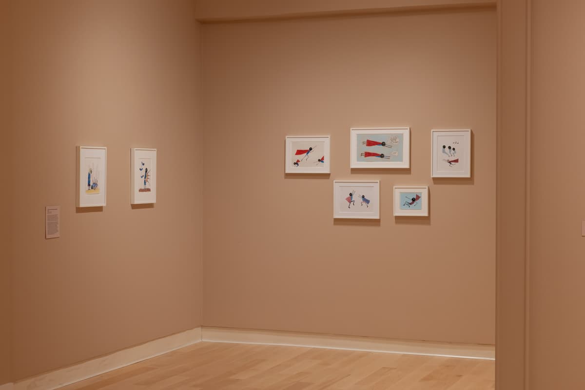 A group of five framed drawings with caped figures adjacent to a wall with two additional framed drawings depicting figures.