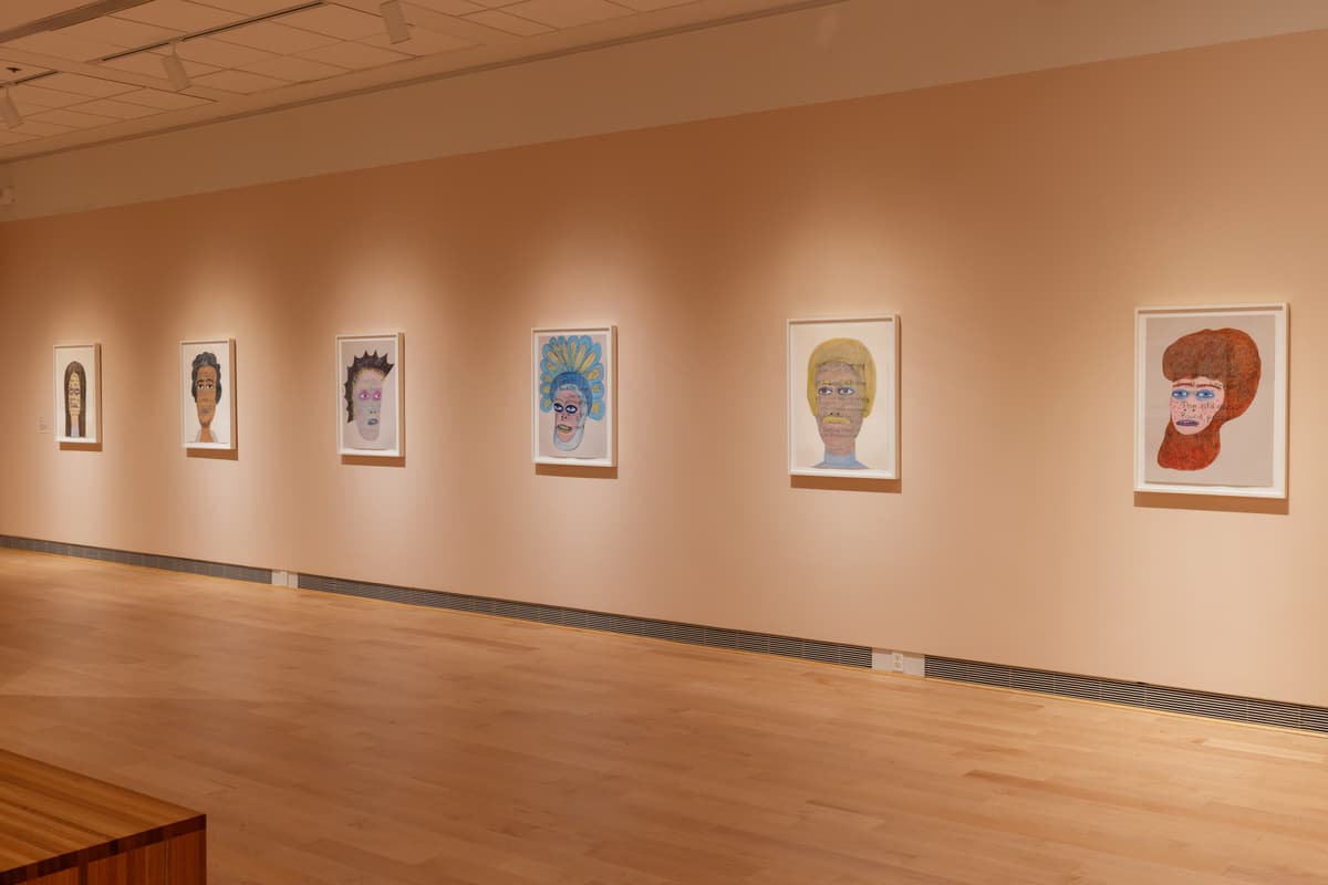Six framed drawings hung in a gallery, each depicting a single figure's face.