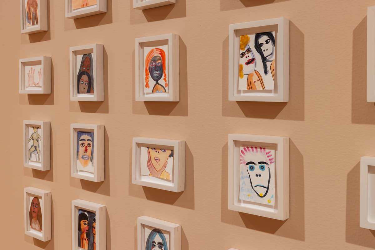 A grid of small, colorful, figurative drawings frames hung on a gallery wall.