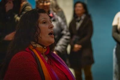 A close-up, candid photograph of a woman with long, dark, curly hair and a red, textured sweater, looking off to the side with her mouth slightly open as if speaking or singing. She is wearing a multicolored scarf (red, orange, purple) and a collar necklace. The background is softly focused, showing a few blurry figures and a muted blue wall, suggesting she is part of an audience or a group presentation in a public space.