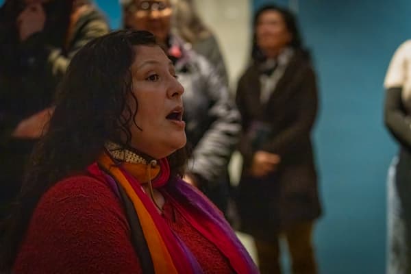A close-up, candid photograph of a woman with long, dark, curly hair and a red, textured sweater, looking off to the side with her mouth slightly open as if speaking or singing. She is wearing a multicolored scarf (red, orange, purple) and a collar necklace. The background is softly focused, showing a few blurry figures and a muted blue wall, suggesting she is part of an audience or a group presentation in a public space.