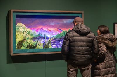 Two people, one wearing a brown hat and black puffer jacket and the other a brown hooded coat, stand in a dimly lit museum gallery looking up at a large, vibrant artwork. The artwork is a detailed, layered paper-cut or mixed-media landscape featuring stylized, jagged pink and purple mountains under a swirling pink and blue sky. The foreground shows dark green and yellow-green forests and plants, creating a striking contrast with the bright, almost neon colors of the mountains and sky