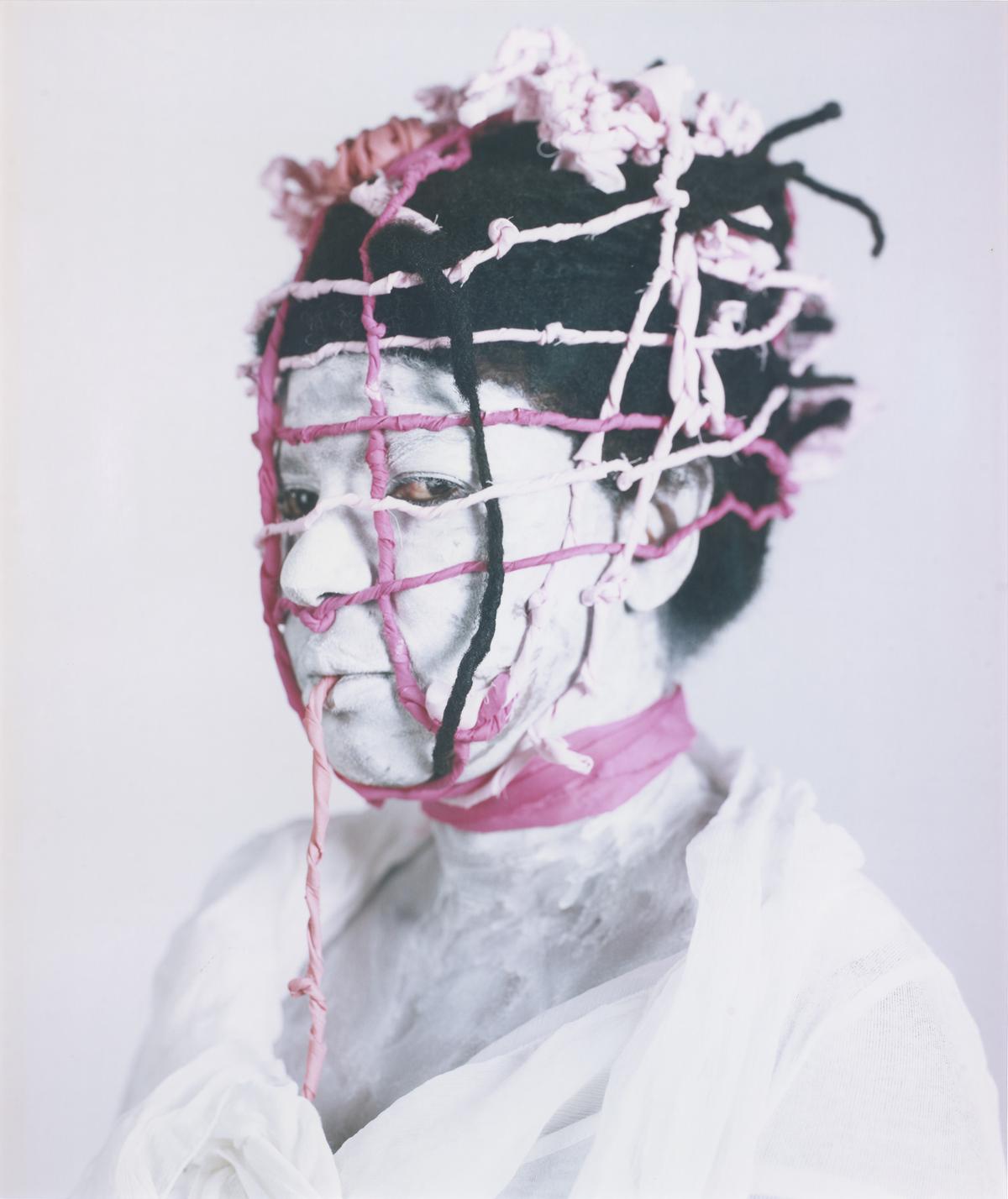 A vertical portrait of a person whose face and neck are covered in white pigment. Their head and face are tightly bound in a grid-like "trap" or cage made of pink and black braided fabric. The subject looks directly at the camera with a neutral expression against a plain white background.
