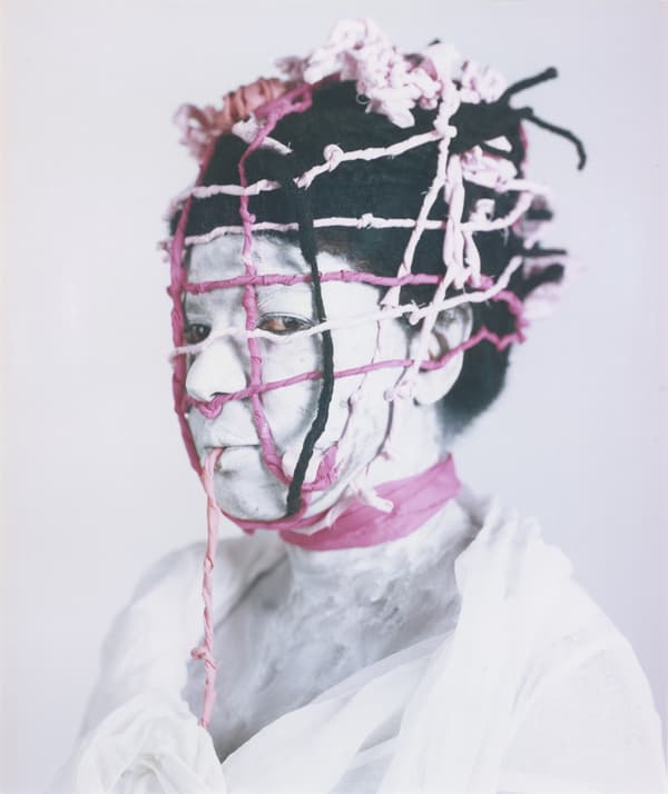 A vertical portrait of a person whose face and neck are covered in white pigment. Their head and face are tightly bound in a grid-like "trap" or cage made of pink and black braided fabric. The subject looks directly at the camera with a neutral expression against a plain white background.