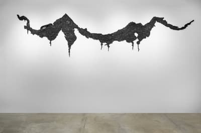 A large-scale wall installation consisting of a long, horizontal, jagged black form that resembles a mountain range or a series of islands seen from a distance. The material appears textured and charcoal-like, with small pieces hanging down like stalactites against a clean white gallery wall.