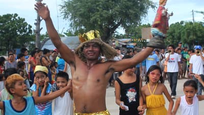 A vibrant film still showing a festival or parade in the Philippines. A man in the center, shirtless and wearing a golden crown and grass-like headpiece, smiles with his arms outstretched. He holds a religious figurine. He is surrounded by a crowd of cheering children and townspeople on a dirt road.