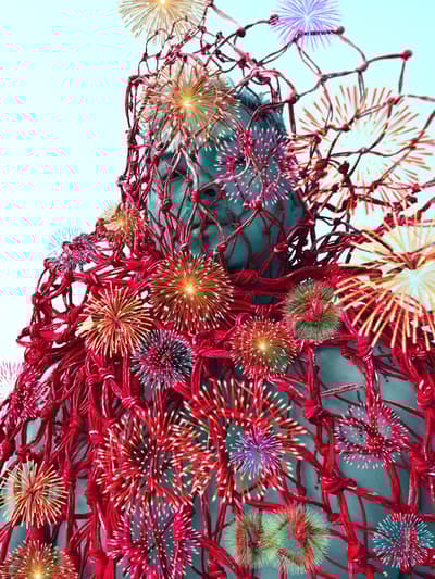 A close-up photograph of a person with blue-tinted skin, looking upward. They are draped in a heavy, bright red fishing net. Digitally overlaid across the image are numerous colorful, glowing starbursts or firework-like patterns in shades of yellow, pink, and green.