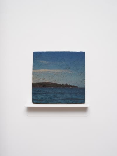 A small, square photographic print mounted on a thick, textured block, which sits on a thin white shelf against a plain white wall. The image depicts a calm blue sea in the foreground and a dark, low-lying landmass or island under a pale blue sky with light clouds. The texture of the block gives the photograph a weathered, stone-like appearance.