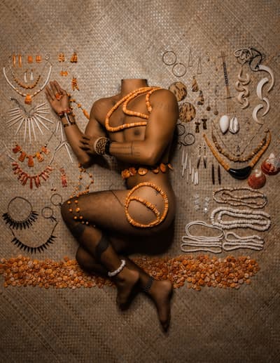 A vertical photograph taken from a high angle, showing a headless, nude person with tanned skin lying in a fetal position on a woven tan mat. The person is adorned with intricate orange shell jewelry across their torso and hip. Surrounding them on the mat is a symmetrical and extensive collection of Indigenous jewelry, including necklaces, earrings, and bracelets made of shells, bone, and dark wood, arranged like a museum display or an anatomical study.