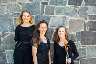 A professional portrait of three women smiling in front of a textured stone wall. They are all dressed in elegant black attire. The woman on the left has long blonde hair and wears a black top with a ribbon belt. The woman in the center has long brown hair and wears a black lace top. The woman on the right has curly reddish-brown hair and holds a cello, with the neck and scroll visible in the foreground. The lighting is bright and natural, casting soft shadows against the grey and brown stones.