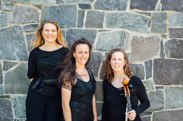 A professional portrait of three women smiling in front of a textured stone wall. They are all dressed in elegant black attire. The woman on the left has long blonde hair and wears a black top with a ribbon belt. The woman in the center has long brown hair and wears a black lace top. The woman on the right has curly reddish-brown hair and holds a cello, with the neck and scroll visible in the foreground. The lighting is bright and natural, casting soft shadows against the grey and brown stones.