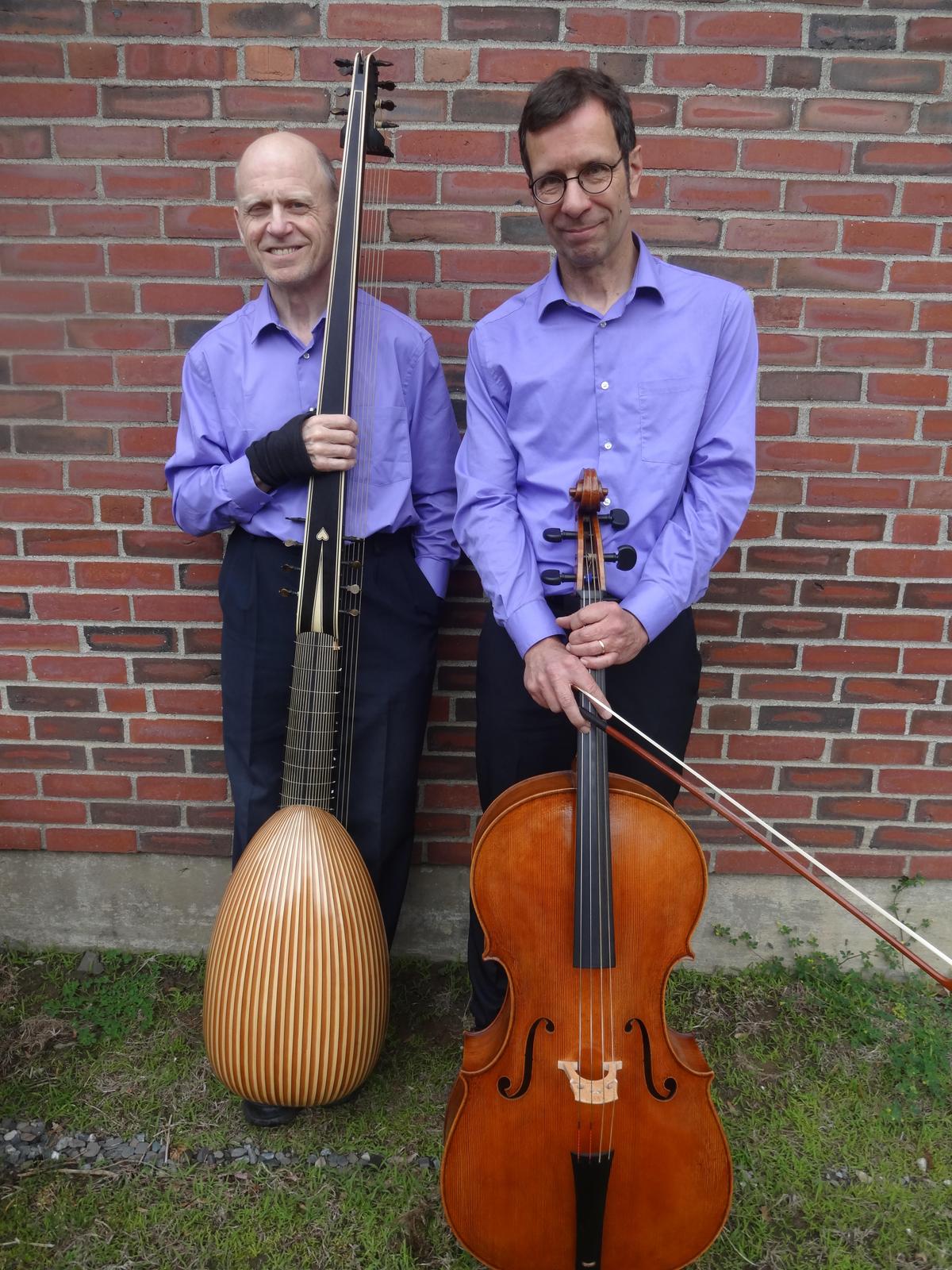 A full-length shot shows two men standing side-by-side against a red brick wall, holding musical instruments. Both men are wearing matching light purple button-down shirts and dark trousers. On the left, a bald man with a smile holds a large theorbo (a long-necked lute) vertically. The instrument has a rounded, striped wooden body and an exceptionally long neck that extends above his head. He has a black support wrap on his right wrist. On the right, a man with short brown hair and glasses stands holding a cello in front of him, with a bow resting diagonally across the strings. They are standing on a patch of grass and dirt in front of the brick building. The lighting is natural and bright.