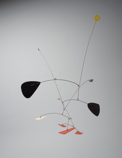 A hanging kinetic mobile sculpture. It consists of thin wire arms supporting various flat, organic, and geometric shapes in black, red, yellow, and white. The piece is suspended against a neutral grey background.