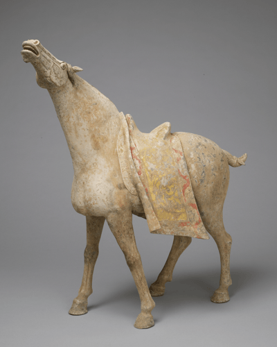 A ceramic sculpture of a horse from the Tang Dynasty. The horse is buff-colored with its head reared back and mouth open in a neigh. It features a decorative saddle blanket with faded red and yellow patterns.