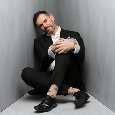 A man with a short beard and dark hair sits in the corner of a grey, minimalist room. He wears a black suit, a white dress shirt with the cuffs unfolded, and tan socks featuring a colorful car pattern. He is seated on the floor with one knee tucked toward his chest, his hands clasped around it, and his head tilted slightly as he looks directly at the camera.