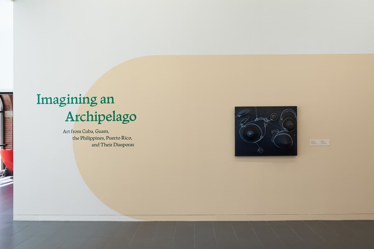 A white gallery wall features a large, light beige arch painted across it. On the left inside the arch, green text reads, "Imagining an Archipelago: Art from Cuba, Guam, the Philippines, Puerto Rico, and Their Diasporas." To the right, a dark square artwork with white line drawings and protruding black cones hangs at eye level alongside two wall labels.