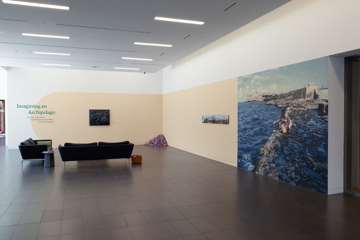 An expansive, brightly lit gallery features a wide beige stripe wrapping around white walls, with dark sofas on grey tiled floors facing artworks. On the far right wall, a large photograph depicts a person sitting on a rocky shore beside deep blue water, while a pile of colorful candies sits in the far corner.