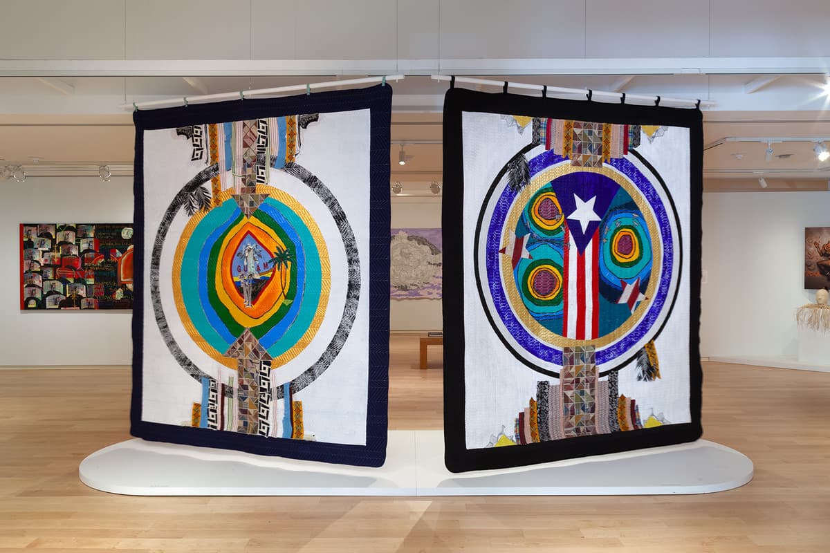 Two large, colorful textiles hang side-by-side from the ceiling above a low white platform in a gallery with light wood floors. Each textile features a white center bordered by dark trim and holds a bold circular emblem incorporating island imagery, including the Guam seal on the left and the Puerto Rican flag on the right.