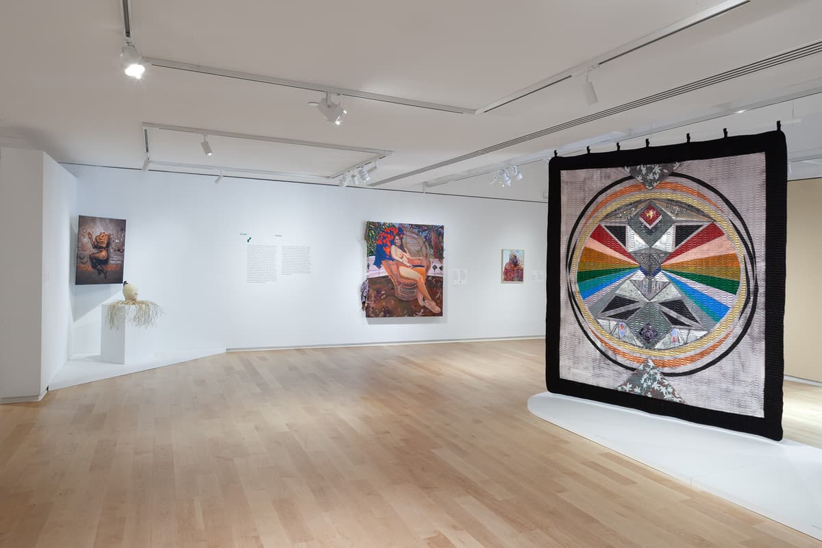 A large textile featuring a vibrant, multi-colored circular geometric design hangs in the foreground of a gallery space with light wood flooring. In the background along white walls, several artworks are displayed, including wall panels with text, mixed-media pieces, and a colorful figurative painting.