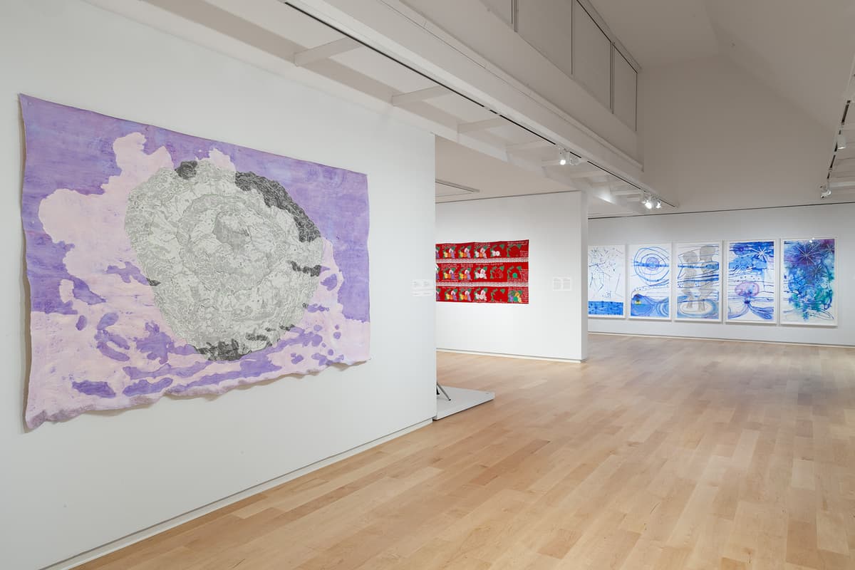 A large wall hanging with a round, grey, map-like shape on a soft purple background anchors the foreground of a gallery with light wood floors. In the adjoining room beyond, a red textile piece and a series of blue, vertical abstract artworks are displayed along white walls.