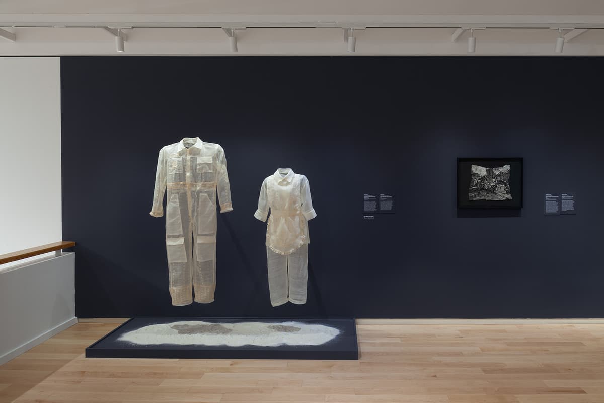 Two semi-transparent white garments—a full-length jumpsuit and a two-piece outfit—hang suspended above a low floor tray filled with sand-like material. They are displayed against a dark navy wall alongside wall text and a framed black-and-white artwork.