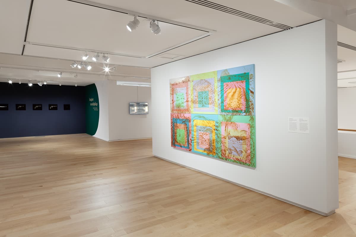 A large artwork composed of six vibrant, pastel-toned panels displaying layered tropical landscapes hangs on a white gallery wall. In the background, light wood floors extend toward adjoining gallery spaces, including a dark wall displaying a row of small, framed images.