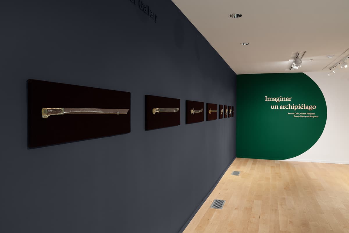 Along a dark grey gallery wall, seven glass sculptures shaped like traditional swords are mounted horizontally in a row on dark plaques. In the background, a white wall features a large dark green arch with text in beige lettering.