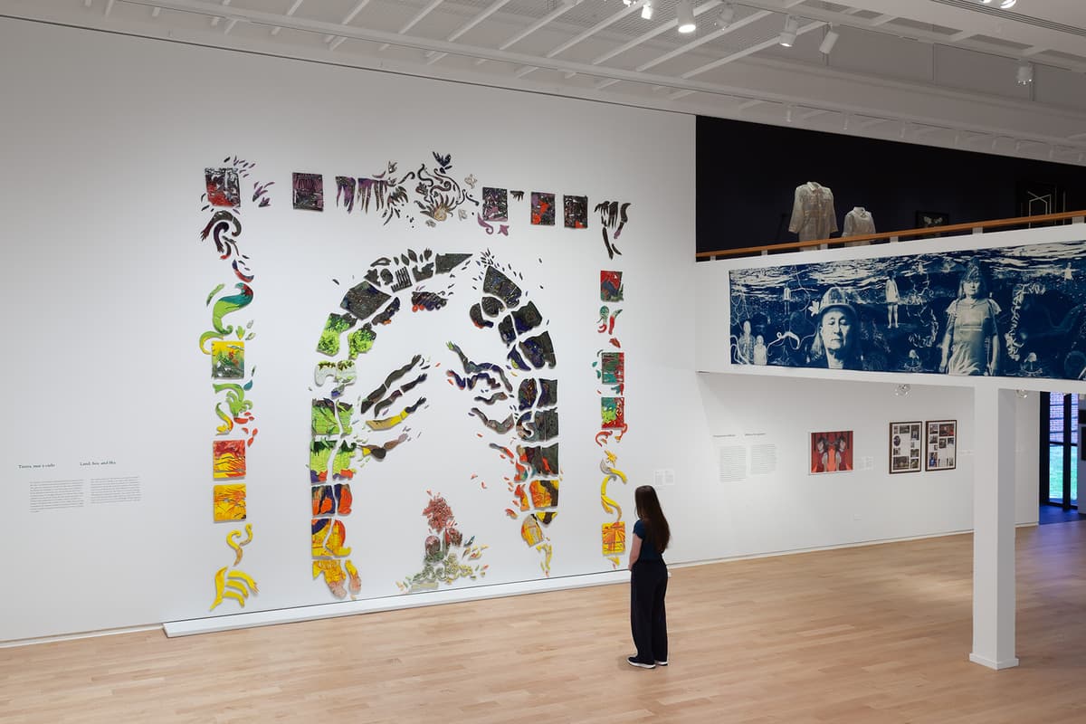 In a high-ceilinged gallery, a large wall installation composed of colorful ceramic tiles forming an arch and surrounding figures fills a white wall. Nearby, a visitor stands observing the display, while a balcony above holds hanging garments and a wide blue-and-white photo mural.