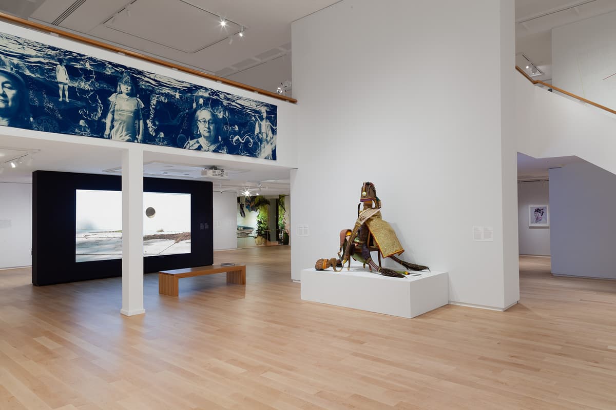 A open gallery with light wood floors displays a complex mixed-media sculpture seated on a low white pedestal. Above, a long blue-toned photographic mural spans an upper balcony wall, while a large video screen built into a black partition rests in the background.
