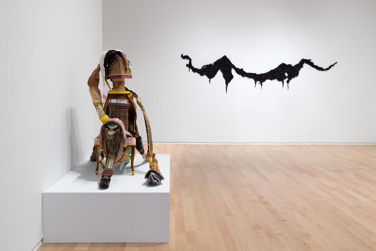 A low white pedestal in a gallery with light wood floors holds an intricate, seated assemblage sculpture crafted from wood, textiles, and found objects. On the white wall behind it, a textured black abstract sculpture stretches horizontally like a mountain range or coastline.