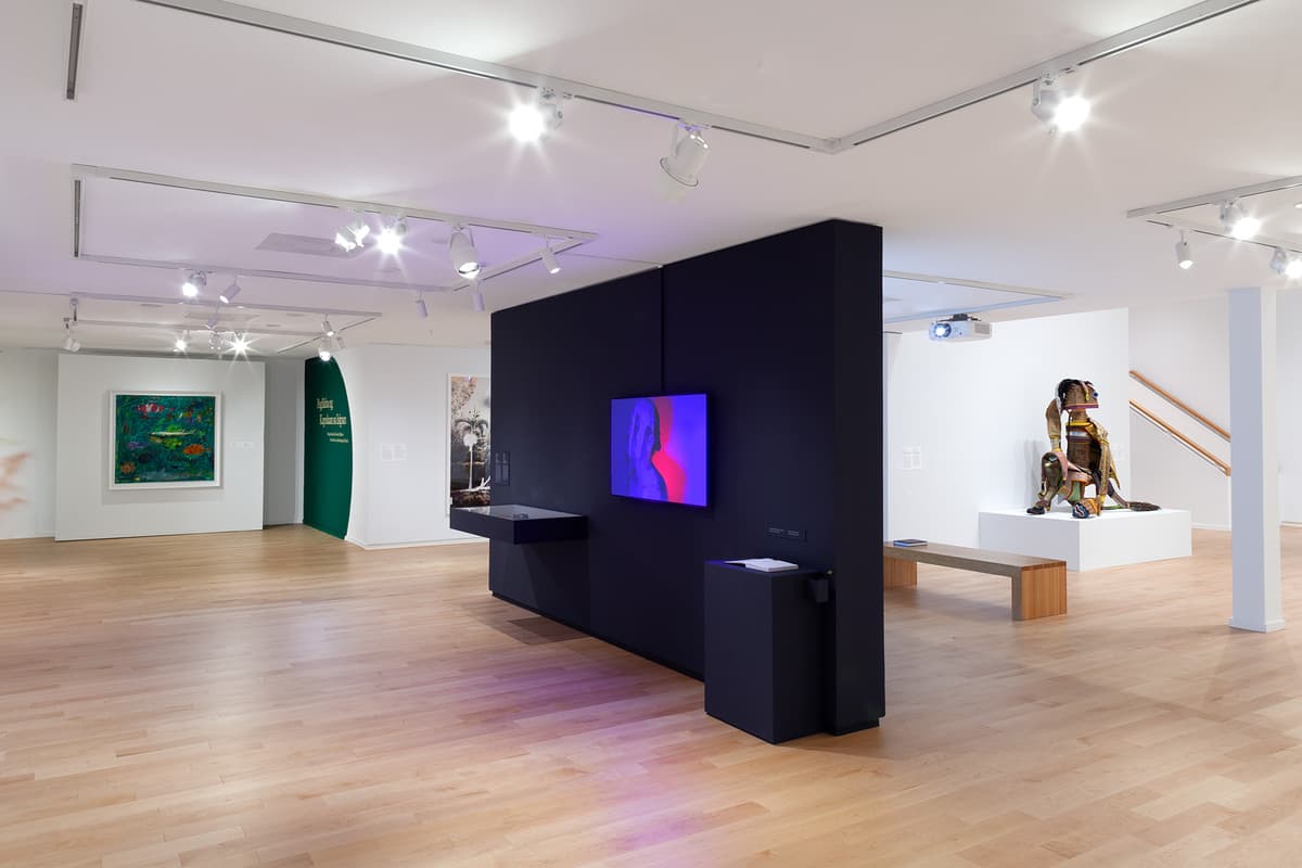 In a spacious gallery with light wood floors, a dark central wall holds a digital screen displaying a portrait in vibrant pink and blue tones. In the background, additional artworks are visible, including a lush green painting, wall text, and a sculpture on a white pedestal.