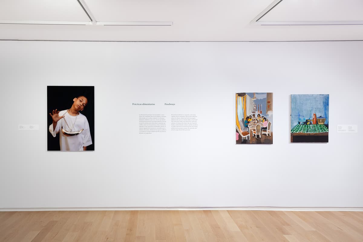 A white gallery wall titled "Prácticas alimentarias / Foodways" displays a large portrait photo of a young boy wearing a gold pendant on the left, and two paintings showing dining and kitchen scenes on the right.
