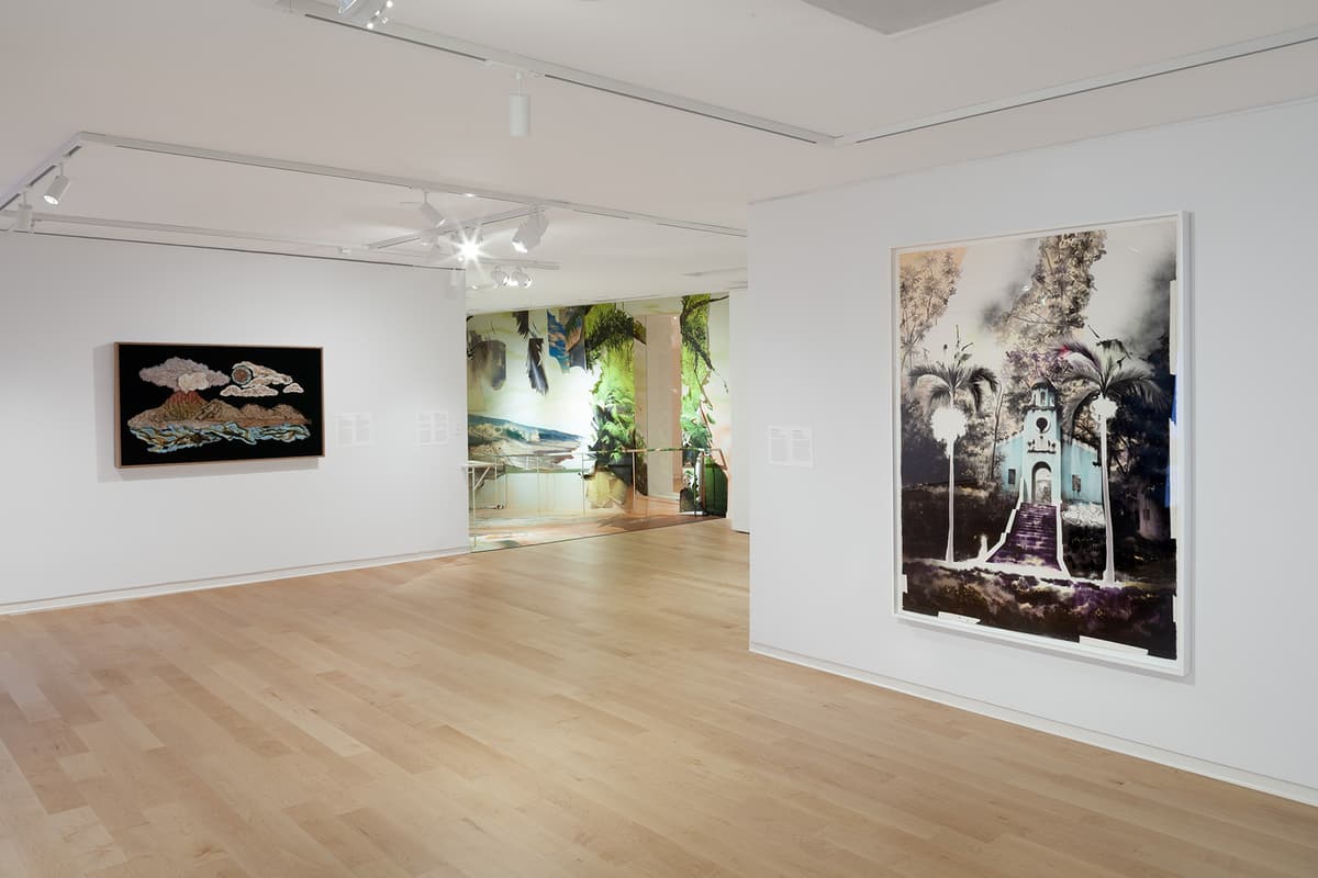 In a gallery space with light wood flooring, a white wall displays a framed painting depicting a blue church framed by palm trees. To the left, a dark embroidered artwork showing a landscape hangs beside an entrance lead-in covered with wallpaper depicting a tropical coastline.