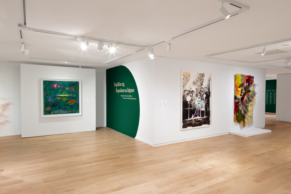A dimly lit gallery wall features an illuminated, dark-toned artwork flanked by text panels. On the left wall, green text titled "Creencias isleñas / Island Beliefs" is displayed, while a small white pedestal with a sculptured object sits nearby on the light wood floor.