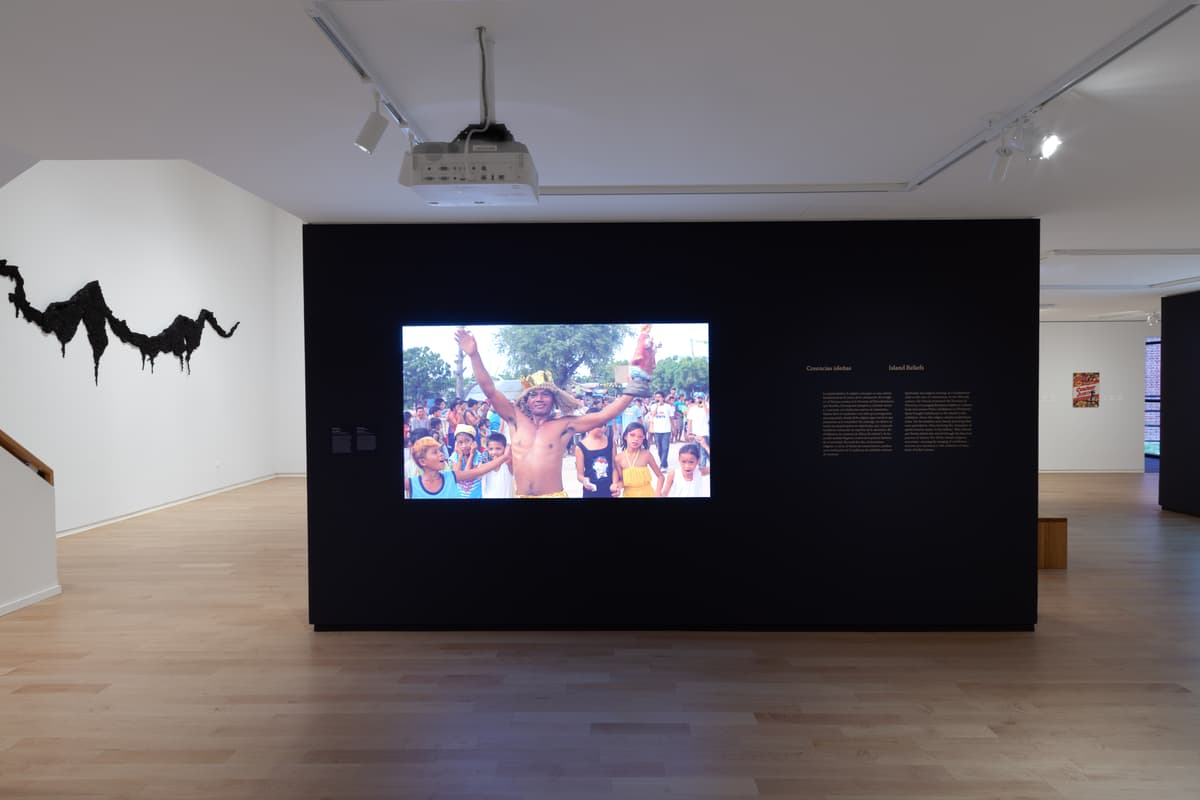 A large video screen mounted on a dark partition projects a scene of a crowd with a man raising his hands in celebration. To the right of the screen, text titled "Creencias isleñas / Island Beliefs" appears on the wall, and an organic black wall sculpture hangs in the background.