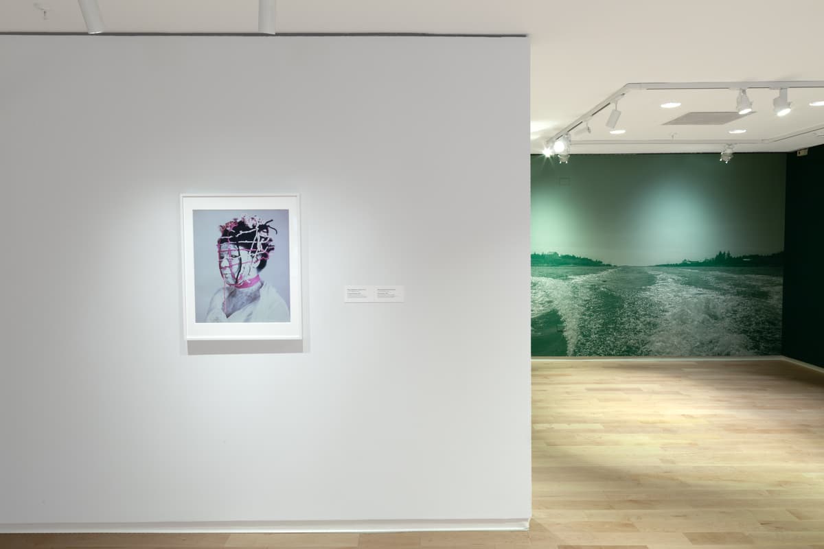 A gallery room featuring a large, dark-toned artwork flanked by bilingual text panels on a white wall. To the left, green wall text titled "Creencias isleñas / Island Beliefs" introduces the section, and a small white pedestal stands near the light wood floor.