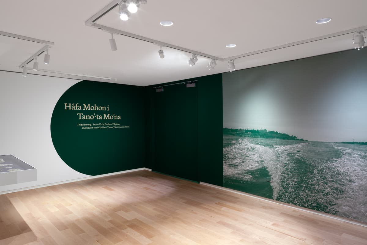 A dark green arch painted on a white wall displays text in Chamorro, leading to an adjacent dark wall with a large black-and-white photograph of a boat's wake on open water. A low display case sits along the far left edge over light wood flooring.
