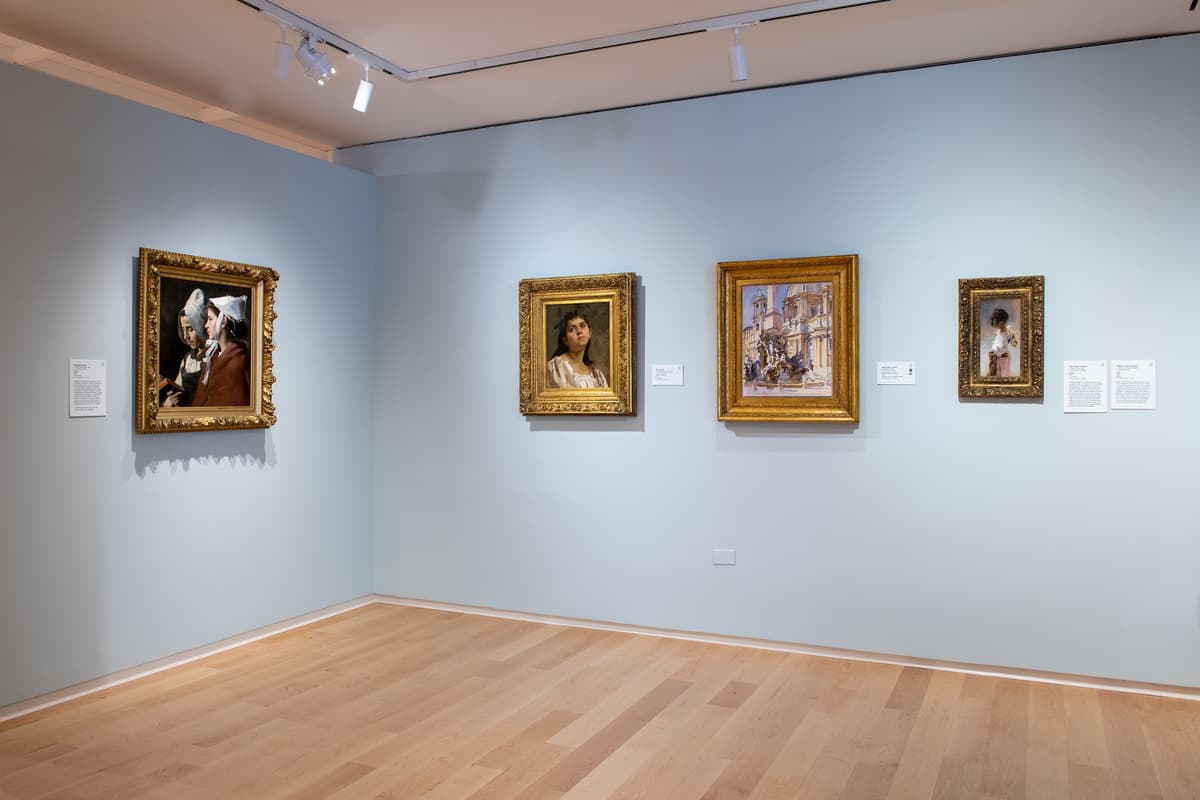 A corner of a gallery with four ornate gold-framed paintings, primarily portraits and a cityscape, illuminated by overhead track lighting against pale blue walls.