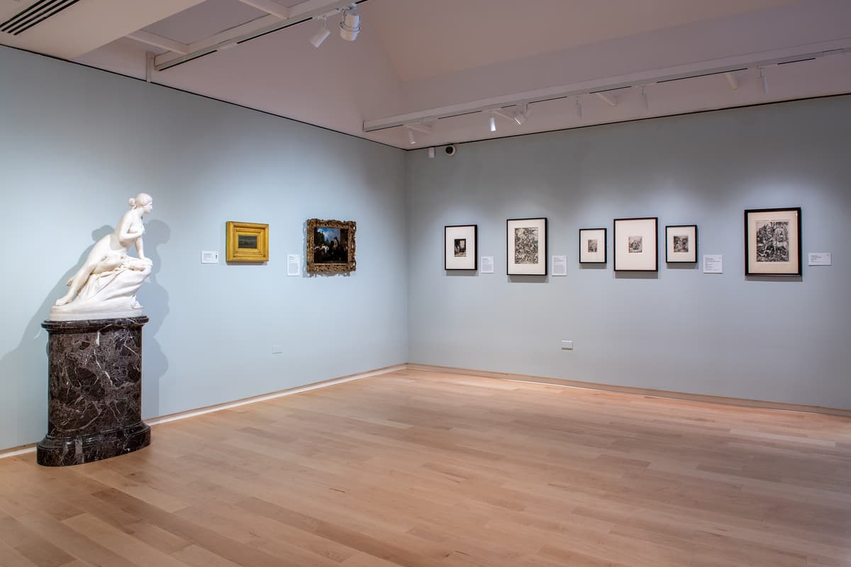 A gallery view showing a white marble sculpture of a reclining woman on a dark stone pedestal, next to a wall with various framed classical sketches and small paintings.