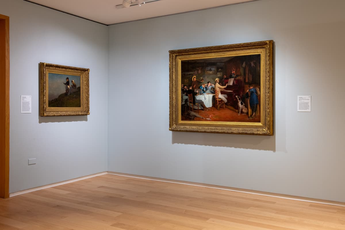 Two framed oil paintings on a light blue wall; the larger painting on the right depicts a lively interior scene with people gathered around a piano.